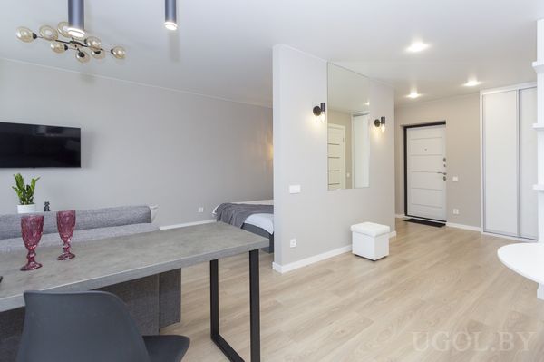 Комната с серым диваном, кроватью, столом с бокалами, телевизором и входной зоной с зеркалом.