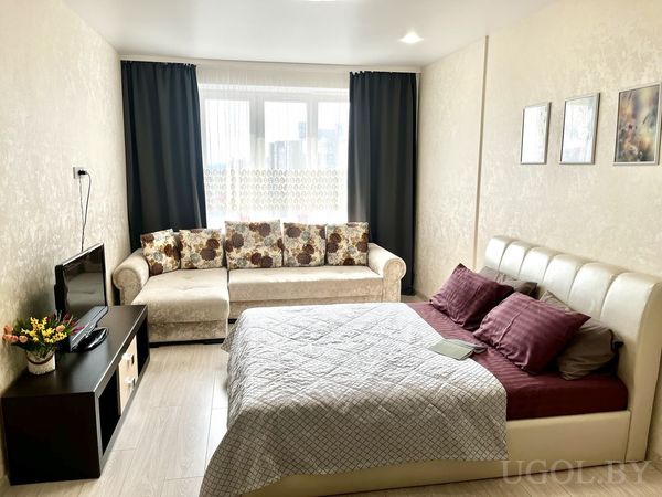 Уютная спальня с белым изголовьем, бордовым покрывалом, телевизором на тумбе и кремовым диваном с подушками.