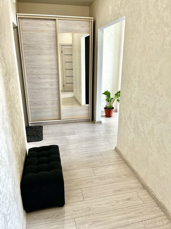 Современный коридор с чёрным пуфом, зеркальным шкафом‑купе и растением в соседней комнате.