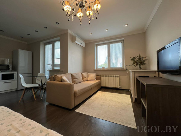 Бежевый диван, белый ковёр, ТВ-зона, люстра и стеклянный стол с белыми стульями на фоне кухни с техникой.