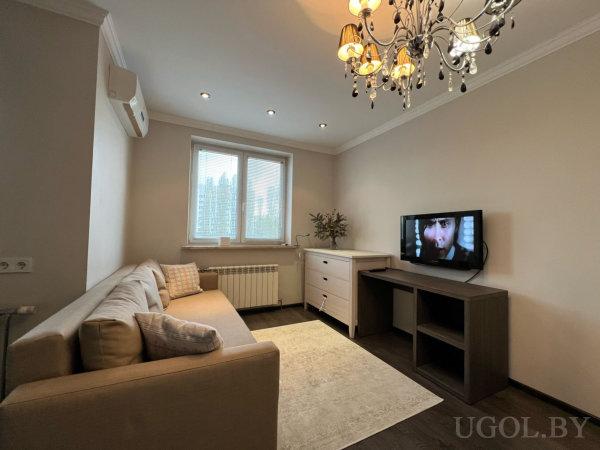 Бежевый угловой диван, настенный телевизор, белая тумба с растением, люстра с подвесами и окно с жалюзи.