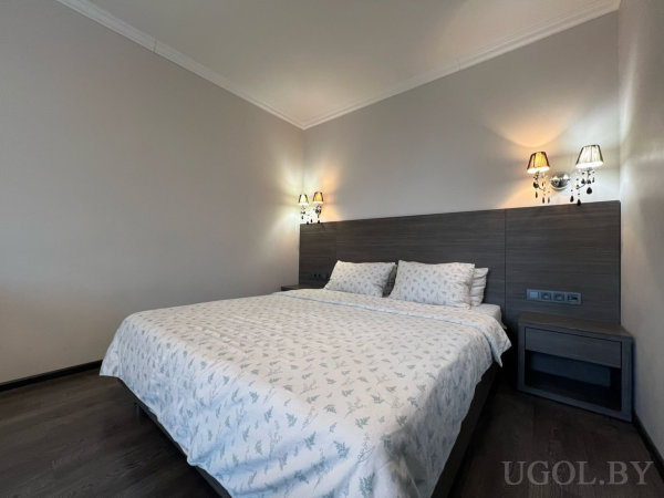 Кровать с белым покрывалом, тёмное изголовье с тумбами, настенные светильники и тёплый свет на фоне бежевых стен.