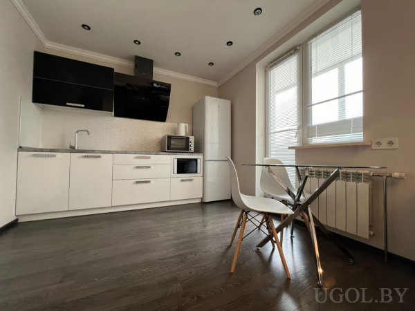 Белые шкафы, варочная панель, микроволновка, холодильник, стеклянный стол с белыми стульями и окно с жалюзи.