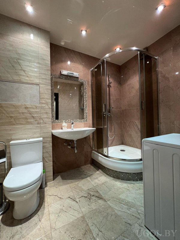 Белая раковина, зеркало с подсветкой, душевая с прозрачными дверцами, унитаз и стиральная машина на фоне бежевой плитки.