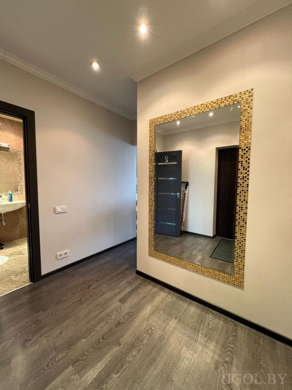 Зеркало с мозаичной рамой, светлый проход, тёмная дверь, точечное освещение и вид на ванную с раковиной.