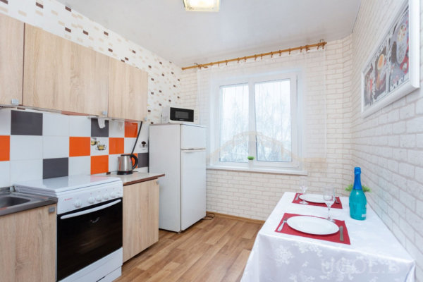 Компактная кухня с деревянными шкафами и плиткой в оранжево-серой гамме, плита и чайник на столешнице, холодильник с микроволновкой сверху. У окна стол с белой скатертью, сервированный на двоих с бокалами и красными салфетками.