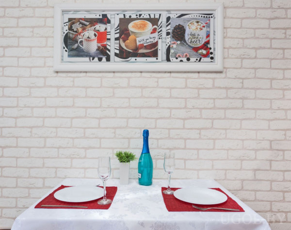 Стол с белой скатертью и красными салфетками, сервированный тарелками и бокалами, в центре синяя бутылка и растение, на стене триптих с изображениями еды и напитков.