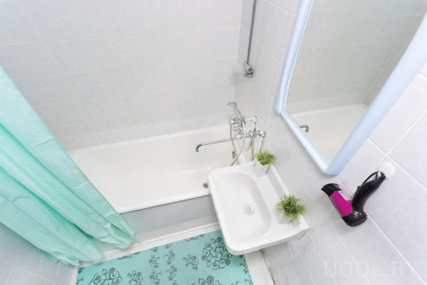 Белая ванна и маленькая раковина с общим смесителем, зеркало над раковиной, фен на стене, два растения для декора, бирюзовый коврик и занавеска с цветочным узором.