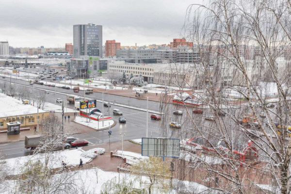 Заснеженные улицы и тротуары, автомобили, автобусы и пешеходы на перекрёстке, фоновые многоэтажные здания и рекламный щит, деревья без листвы создают зимнюю атмосферу.