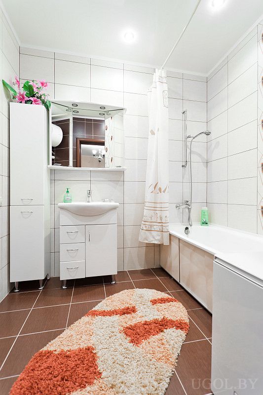 Чистая ванная с белой мебелью, зеркальным шкафом, душем и ярким ковриком. Есть высокий пенал и декоративные цветы.