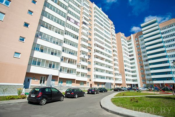 Современный жилой комплекс с парковкой, зелёной зоной и детской площадкой. Чистый фасад, остеклённые балконы и солнечная погода.