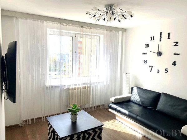 Комната с диваном, столом, телевизором, настенными часами, торшером и тюлем.