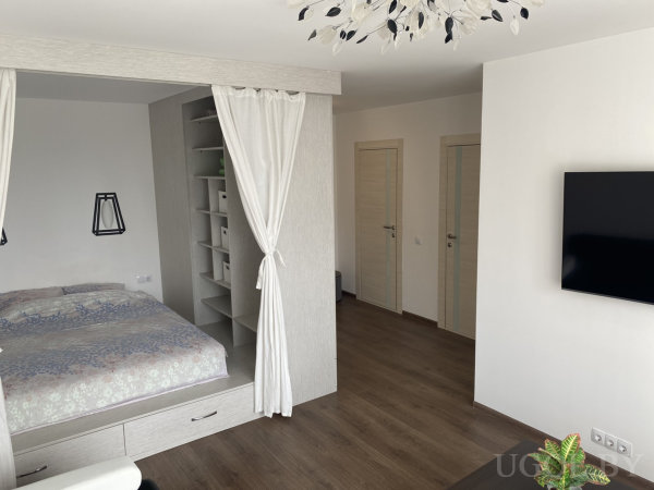 Интерьер с кроватью, полками, шторой, светильником, телевизором и деревянным полом.