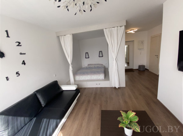 Интерьер с кроватью, диваном, столом, телевизором, часами и проходом в коридор.