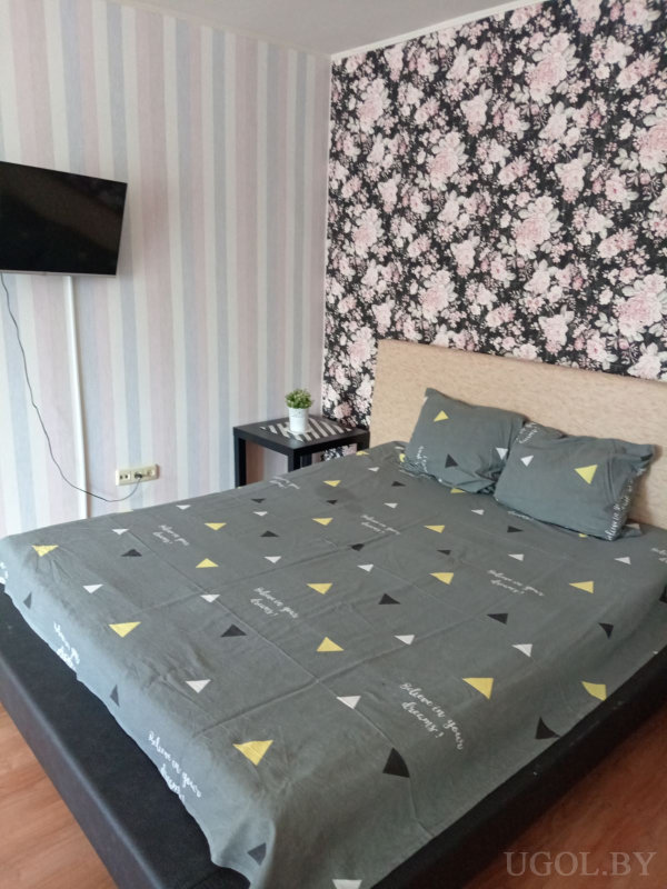 Кровать с серым покрывалом и узором из треугольников, бежевое изголовье, стена с чёрными обоями и крупными розовыми цветами, рядом тумбочка с растением, напротив телевизор на полосатой стене.