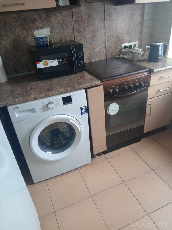 Белая стиральная машина «beko», рядом микроволновка и коричневая плита с трещиной на стекле, электрический чайник и кружки на столешнице, коричневые шкафы и плитка в тон.