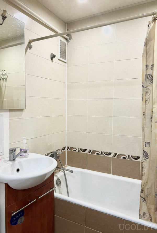 Белая ванна с душем и шторой с узором ракушек, рядом раковина с деревянной тумбой и дозатором, над ней зеркало и вентиляционная решётка, стены в бежево-коричневой плитке, компактный и аккуратный интерьер.