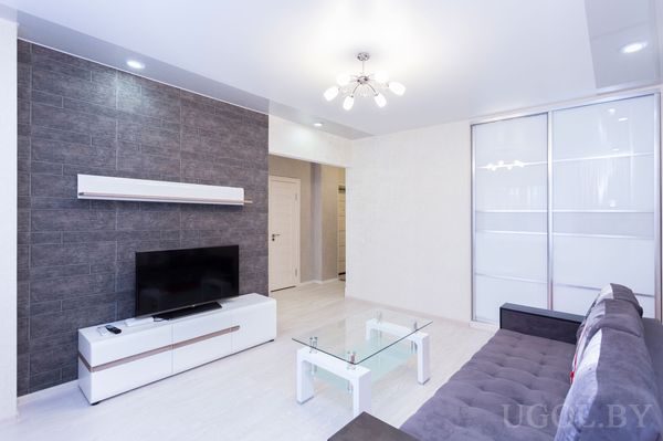 Комната с диваном, телевизором, белой тумбой, стеклянным столом, зеркальным шкафом и подвесным светильником.