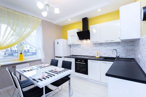 Интерьер с белыми фасадами, чёрной техникой, жёлтым чайником, вазой, тюлем и обеденной зоной.