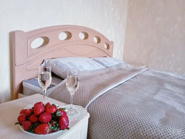 Аккуратно застеленная кровать с серым покрывалом, на прикроватном столике тарелка клубники и два бокала.