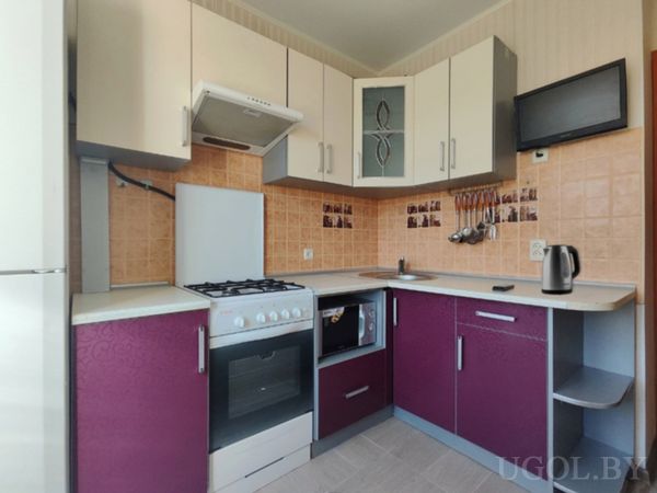 Современная кухня с бело-фиолетовыми шкафами, плитой, вытяжкой и настенным телевизором.