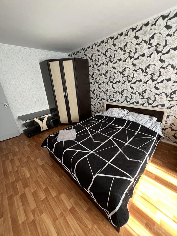 Кровать с чёрным покрывалом, подушками в цветочном узоре, двухцветной мебелью и солнечным светом на полу.