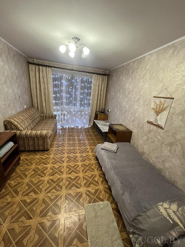 Уютная комната с серой кроватью, диваном в полоску, столом у окна и гирляндой на бежевых шторах.