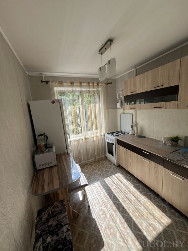 Кухня с белым холодильником, плитой, вытяжкой, деревянными фасадами и столом у окна с лавкой.