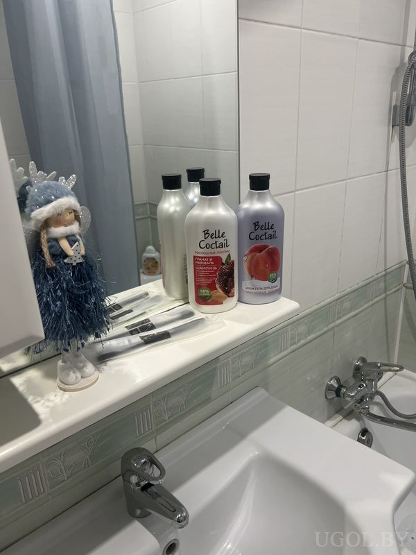 Полка с шампунями Belle Coctail, зубными щётками и куклой в зимнем наряде на фоне белой плитки.