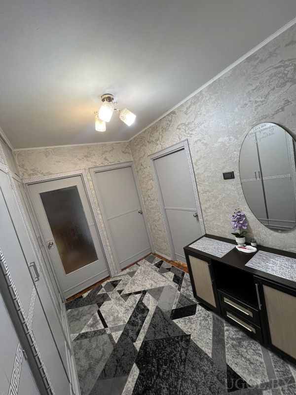 Коридор с серыми дверями, ковром в геометричном узоре, тумбой с декором и овальным зеркалом.