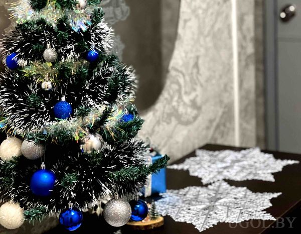 Новогодняя ёлка с синими и серебристыми шарами, подарками и кружевной подложкой на фоне серой стены.