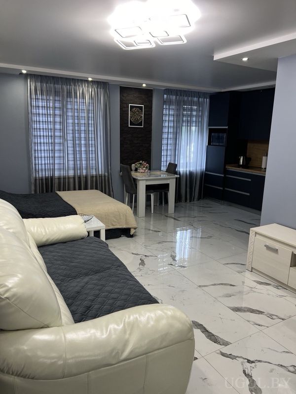 Современная студия с белым диваном, кроватью с бежевым покрывалом, кухней с тёмными фасадами и мраморным полом.