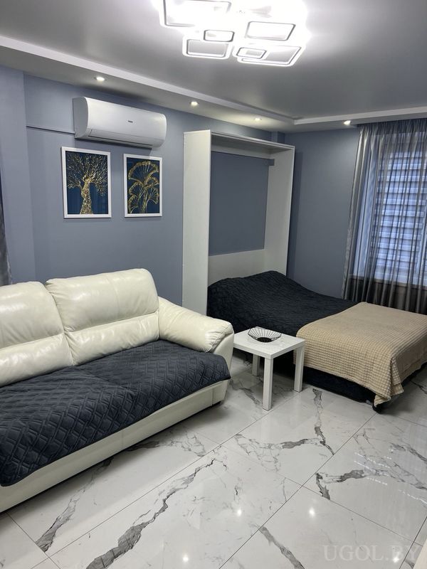 Уютная комната с белым диваном, кроватью с тёмным текстилем, картинами с деревьями и кондиционером над ними.