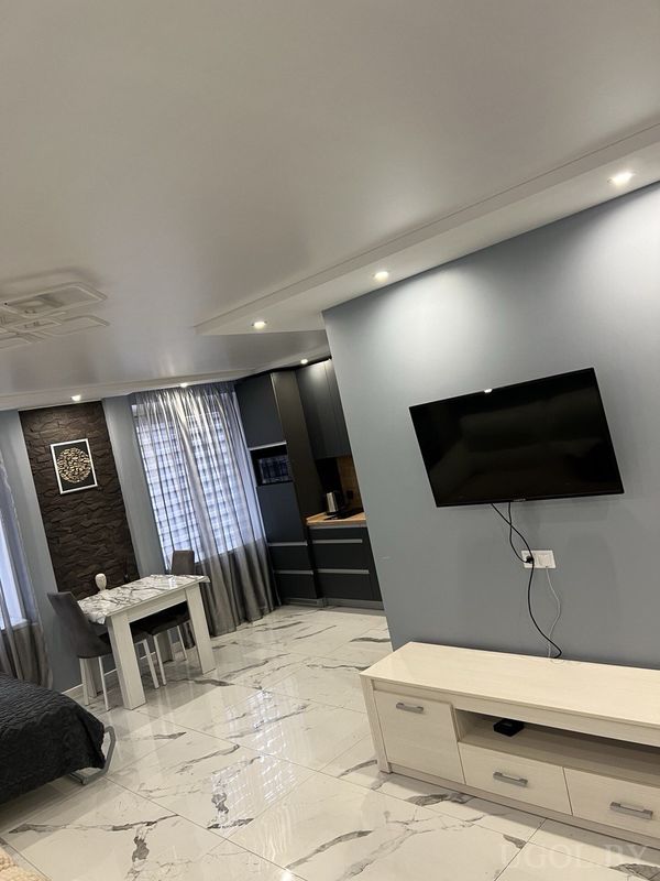 Светлая студия с телевизором на голубой стене, обеденной зоной у окна и кухней с тёмными фасадами.