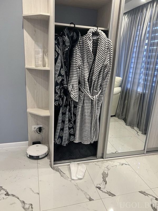 Угловая зона с открытым шкафом, двумя халатами, белыми тапочками и роботом-пылесосом на мраморном полу.