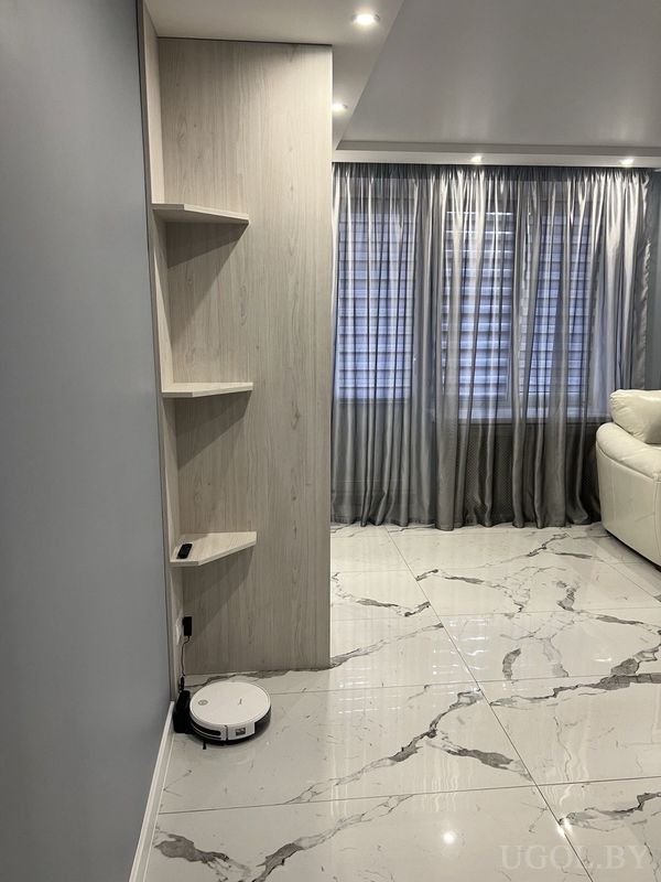 Минималистичная гостиная с белым диваном, роботом-пылесосом, деревянными полками и серыми шторами у окна.