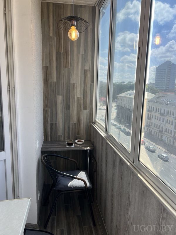Компактный балкон с деревянной отделкой, чёрным креслом, подвесным светильником и видом на городские дома.