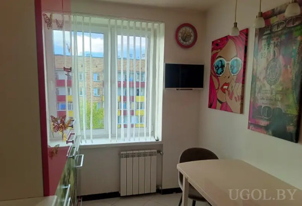 Небольшая столовая с белым столом, телевизором, часами и картинами в стиле поп-арт, включая женщину с розовыми волосами.