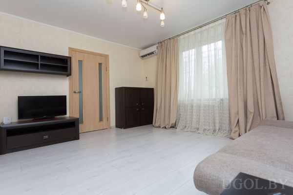 Светлая гостиная с телевизором на тумбе, настенными полками, большим окном и бежевыми шторами.