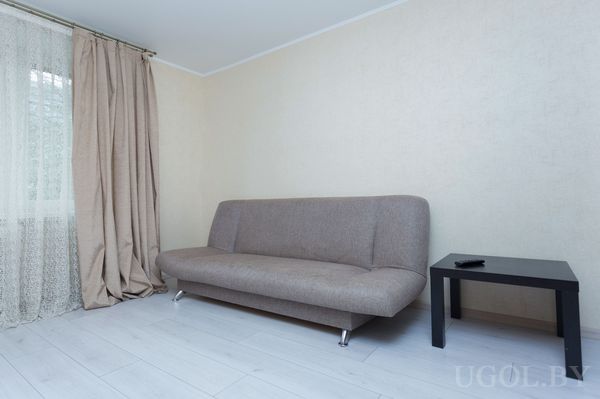 Комната с бежевым диваном, чёрным столиком, окном с белыми шторами и спокойным нейтральным интерьером.