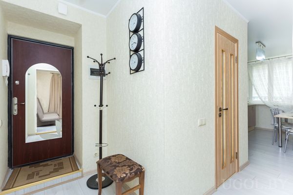 Светлая прихожая с зеркальной дверью, вешалкой, пуфом и часами на стене.