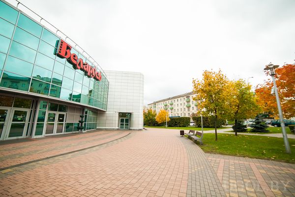 Современное здание с витражным фасадом и вывеской Белмаркет, окружённое деревьями и скамейками.