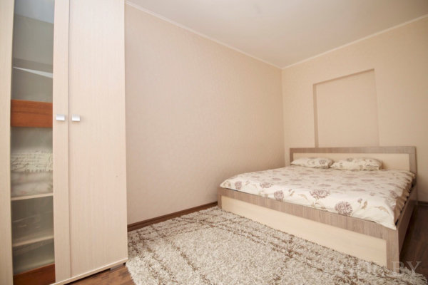 Кровать с цветочным покрывалом и светлым каркасом, рядом шкаф с открытой дверцей и полками, на полу ковёр бежевого оттенка, стены светлые с декоративной нишей над изголовьем.