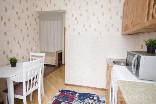 Белый круглый стол с двумя стульями и растением, кухня с мойкой, микроволновкой и стиральной машиной, деревянные шкафы и светлый ламинат, на стене плитка с узором, в соседней комнате видна кровать.