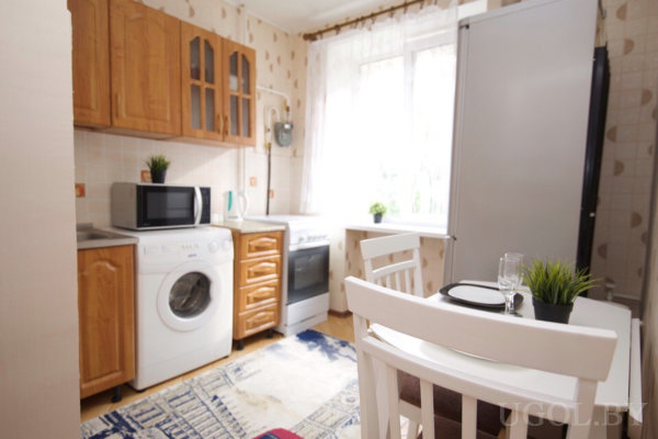 Компактная кухня с деревянными шкафами, плитой, холодильником и стиральной машиной, рядом небольшой стол с белыми стульями и сервировкой, большое окно с тюлем и коврик на полу.