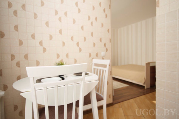 Белый круглый стол с двумя стульями и сервировкой, на столе декоративное растение, стены с плиткой в геометрическом узоре, пол светлый, в соседней комнате видна кровать с бежевым покрывалом.