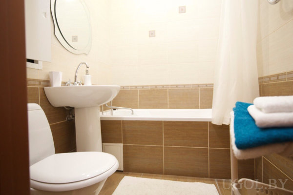 Белый унитаз и раковина с хромированным смесителем, над ней круглое зеркало, рядом ванна с белой шторой, на стене полотенцесушитель с аккуратно сложенными полотенцами, интерьер в бежево-коричневой гамме.