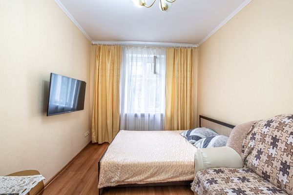 Небольшая спальня с двуспальной кроватью, клетчатым диваном, телевизором на стене и окном с жёлтыми шторами.