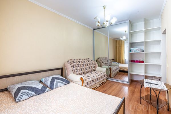 Светлая спальня с двуспальной кроватью, клетчатым диваном, зеркальным шкафом‑купе и полками с вещами.