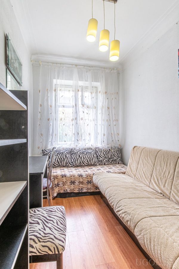 Узкая комната с двумя диванами, рабочим столом с креслом в зебровом стиле и окном с белыми шторами.